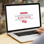 Sobre a mesa, um notebook apresentando a seguinte mensagem: Black Friday 2020. À direita da imagem, a mão de uma pessoa usando o teclado do notebook.